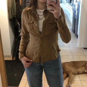 Brun mocka skjorta från H&M - Säljer en brun figursydd mocka skjorta från H&M med klassisk krage och knappar framtill. Skjortan har två bröstfickor med lock och långa ärmar. I strl 36