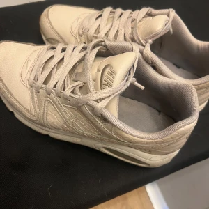 Beige Nike Air sneakers - Säljer ett par beige Nike Air sneakers med klassisk design och synlig luftdämpning i sulan. Skorna har snörning och är tillverkade i syntetmaterial med textilfoder. Perfekta för dig som gillar stilrena och bekväma sneakers.