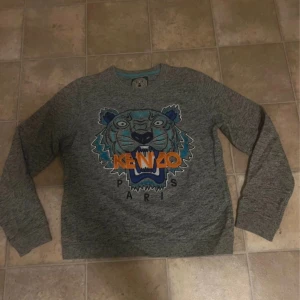 Kenzo Paris tröja - Säljer en grå sweatshirt från Kenzo med tigerbroderi och logga i blått och orange framtill. Tröjan är långärmad och tillverkad i 100% bomull. Tröjan är väldigt sparsamt använd
