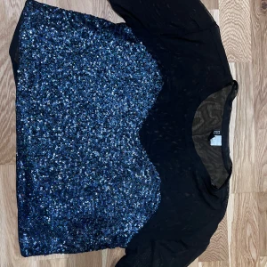 Svart topp med blå paljetter från Divided - Säljer en svart topp från Divided med massor av blå paljetter framtill. Toppen har rund halsringning och korta ärmar. Perfekt för dig som vill sticka ut med lite extra glitter.  Är helt ny, dock utan prislapp.  Kommer från djurfritt hem!