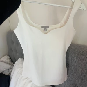 Vit topp från H&M - Säljer en vit topp från H&M med breda axelband och snygg, något hjärtformad urringning. Toppen är enkel och passar perfekt till sommaren. Materialet känns mjukt och stretchigt.