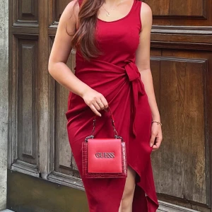Röd omlottklänning och Guess väska  - Snygg röd omlottklänning med knyt i midjan. Klänningen är ärmlös och har en asymmetrisk slits framtill. Perfekt för dig som vill sticka ut med en elegant och modern look. Guess väskan säljs separat. Skriv för mer info❤️