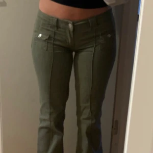 Olivgröna byxor med fickdetaljer - Säljer ett par olivgröna byxor med utsvängda ben och fickor framtill med knappar. Byxorna har normal passform och är lowwaist. Snygga sömmar längs benen och klassisk look. Perfekta till många olika outfits!