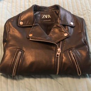 Svart skinnjacka från Zara - Snygg svart skinnjacka från Zara med dragkedjor och knappar i silver. Jackan har en klassisk bikerstil med bälte i midjan och flera dragkedjefickor. Perfekt för en cool look!