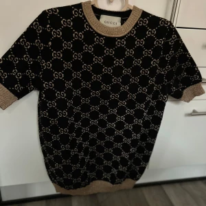 Svart och guld tröja från Gucci - Snygg svart tröja från Gucci med korta ärmar och ett elegant mönster i guld. Tröjan har ribbade detaljer vid hals, ärmslut och nederkant. Perfekt för en stilren look.