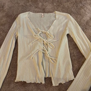 Vit ribbad tröja med knyt - Säljer en stilren vit ribbad tröja med v-ringning och knyt framtill. Tröjan har långa ärmar och en elegant design som passar perfekt för olika tillfällen.