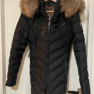 Svart dunjacka med pälsdetalj - Snygg svart dunjacka med dragkedja och pälsdetalj på huvan. Jackan har ett leopardmönstrat foder och flera praktiska fickor med dragkedjor. Perfekt för kalla dagar.