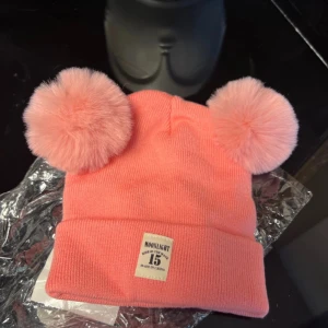 Rosa mössa med pälstofsar - Söt rosa mössa med två stora pälstofsar på toppen. Mössan har en etikett framtill och är perfekt för att hålla värmen under kyliga dagar.