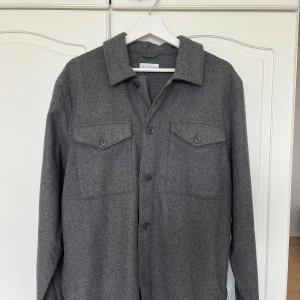 Grå overshirt från These Glory Days - Säljer en stilren grå overshirt från These Glory Days. Den har en klassisk krage och knäppning framtill. Två bröstfickor med knappar ger en snygg detalj. Perfekt för lager-på-lager-stil.