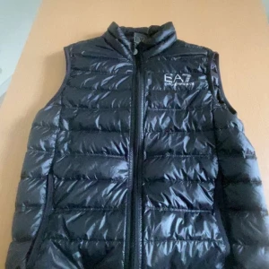 Svart dunväst från EA7 - Snygg svart dunväst från EA7 med dragkedja framtill. Perfekt för kyligare dagar och ger en sportig look. Västen har en hög krage och är quiltad för extra värme.