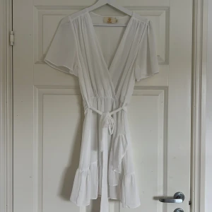 Vit omlottklänning från Chelsea - Säljer en söt vit omlottklänning från Chelsea i storlek XS. Klänningen har korta ärmar och en v-ringad design med knytband i midjan. Den har volanger i nederkanten som ger en fin touch. Perfekt för sommarens alla tillfällen eller likaså konfirmation och student !❤️använd 1 gång sedan tidigare o är som ny!