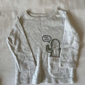 Grå långärmad tröja från Carter's - Söt grå långärmad tröja från Carter's med en kaktus som säger 'Free Hugs'. Perfekt för små barn, tillverkad i mjukt material för bekvämlighet. Tröjan har en liten ficka framtill där kaktusen är placerad.