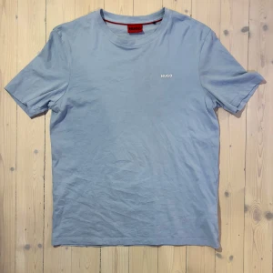 Ljusblå t-shirt från Hugo - Säljer en stilren ljusblå t-shirt från Hugo med korta ärmar och en liten logga på bröstet. Perfekt för en avslappnad look. T-shirten är gjord i mjukt bomullsmaterial.