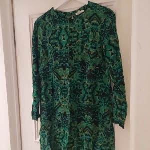 Grön mönstrad klänning - Säljer en snygg grön klänning med ett unikt mönster i svart och guld. Klänningen har trekvartsärmar och en rund halsringning. Perfekt för att sticka ut med stil!