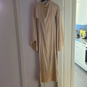 Champagnefärgad långklänning - Elegant champagnefärgad långklänning med långa ärmar och en vacker draperad halsringning. Klänningen har en knytning i nacken som ger en stilfull detalj. Perfekt för speciella tillfällen.