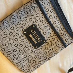 Beige och svart axelväska från Guess - Axelväska från Guess i beige och svart med ett monogrammönster. Väskan har en justerbar axelrem och en dragkedja upptill. Har aldrig använt. 