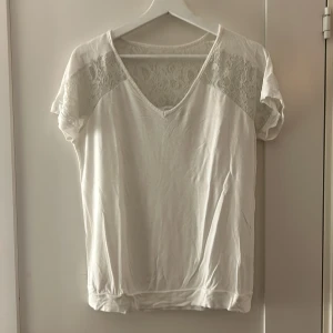 Vit blus från Next - Säljer en stilren vit blus från Next med v-ringning och spetsdetaljer på axlarna och övre delen av ryggen. Blusen har korta ärmar och en lös passform, perfekt för en avslappnad look. Den har ett litet hål på den högra axeln. Passar bra till både jeans och kjol. 🌼