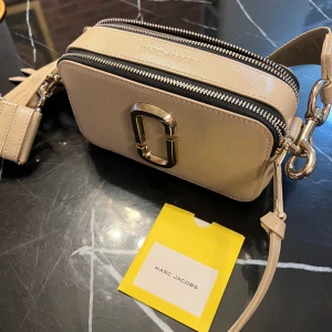 Beige axelväska från Marc Jacobs - Snygg beige axelväska från Marc Jacobs med en stilren design. Väskan har en justerbar axelrem och en praktisk dragkedja. Den är perfekt för att bära dina nödvändigheter med stil. Den har även en dekorativ metallogo på framsidan. ((ÄKTA))