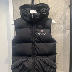 Svart dunväst från Kingsland - Snygg svart dunväst från Kingsland med dragkedja och huva. Perfekt för kyliga dagar  i stallet och har en stilren design med broderad logga på bröstet. Västen är en del av Kingslands Classic Collection.