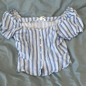 Randig blus från H&M - Säljer en söt blus från H&M i ljusblått och vitt med randigt mönster. Blusen har korta puffärmar och knappar framtill. Den har en volangdetalj vid nederkanten som ger en extra touch. Perfekt för en somrig look! 🌸