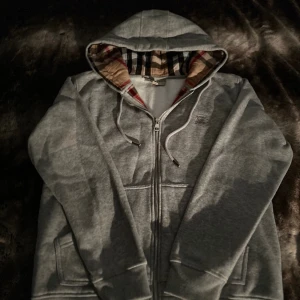 Grå huvtröja med dragkedja - Säljer en grå huvtröja från Burberry med dragkedja och klassiskt rutigt mönster i huvan. Tröjan har långa ärmar och ribbade muddar. Perfekt för en avslappnad stil.