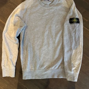 Grå sweatshirt från Stone Island - Säljer en stilren grå sweatshirt från Stone Island med deras ikoniska märke på ärmen. Tröjan har långa ärmar och en klassisk rund halsringning. Perfekt för en avslappnad och trendig look.