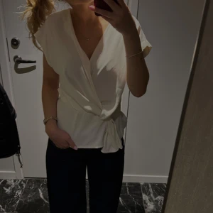 Vit omlottblus - Säljer en stilren vit omlottblus med korta ärmar och knytning i midjan. Perfekt för en chic och avslappnad look. Endast använd en gång men sömmen på ena ärmen har lossnat men det är bra att sy fast igen! 
