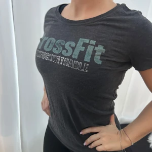 Mörkgrå CrossFit t-shirt - Säljer en mörkgrå t-shirt från CrossFit med texten 'UNFUCKWITHABLE' i ljusblått och vitt. T-shirten har en normal passform och korta ärmar, perfekt för träning eller vardagsbruk.🩶🩵