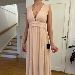 Champagnefärgad långklänning - Elegant champagnefärgad långklänning med djup v-ringning och breda axelband. Klänningen har en vacker drapering och markerad midja som ger en smickrande silhuett. Perfekt för speciella tillfällen.