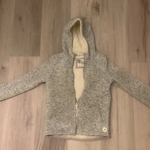 Grå fleecejacka med huva - Mysig grå fleecejacka med dragkedja och huva. Insidan är fodrad med mjuk teddyfleece för extra värme. Perfekt för kyliga dagar. (134/140) i size. 