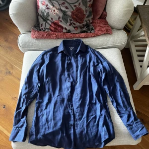 Blå linneskjorta från Massimo Dutti - Snygg blå skjorta i 100% linne från Massimo Dutti. Perfekt för en stilren look med klassiska knappar framtill och långärmad design. Skjortan har en avslappnad passform och är tillverkad i ett lätt och luftigt material.