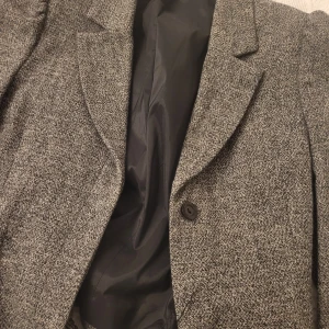 Grå kavaj med dekorativa detaljer - Snygg grå kavaj med ett klassiskt snitt och dekorativa knappar längs kanten. Perfekt för att ge en stilren touch till din outfit. Kavajen har en knapp framtill och är fodrad med ett mörkt tyg.