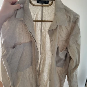 Beige skjorta från Dr. Basic - Snygg beige skjorta från Dr. Basic med klassisk krage och knappar framtill. Skjortan har två bröstfickor och är tillverkad i ett lätt och luftigt material, perfekt för varmare dagar.