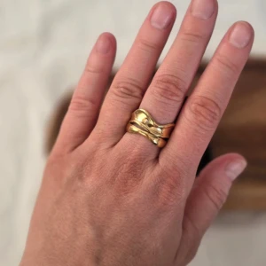Guldfärgad ring med vågig design - Elegant guldfärgad ring med en unik vågig design. Ringen har en bred form som ger ett stilrent och modernt intryck. Perfekt för att ge en extra touch till din outfit.