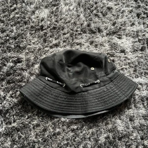 Svart bucket hat med dragsko - Snygg svart bucket hat med justerbar dragsko och små ventilationshål. Perfekt för en avslappnad stil.
