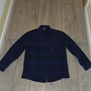 Mörkblå rutig overshirt från H&M - Snygg mörkblå rutig overshirt från H&M i regular fit. Skjortan har långa ärmar och knappar framtill. Perfekt för en stilren look. Oanvänd.