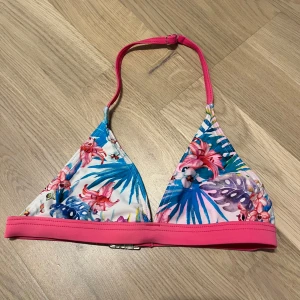 Blommig bikiniöverdel med rosa detaljer - Snygg bikiniöverdel med tropiskt blommönster i blått och rosa. Den har en rosa kant och knytband runt nacken. Perfekt för sommarens stranddagar!