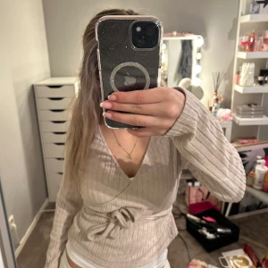Beige ribbad omlottopp - Säljer en snygg beige ribbad omlottopp med långa ärmar och knytband i midjan. Perfekt för en stilren look. Passar bra till både jeans och kjol.