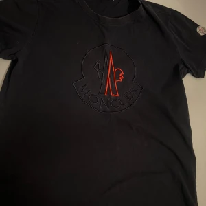 Svart t-shirt från Moncler - Snygg svart t-shirt från Moncler med ett stort broderat märke i rött och svart på framsidan. T-shirten har korta ärmar och en rund halsringning. Perfekt för en stilren look.