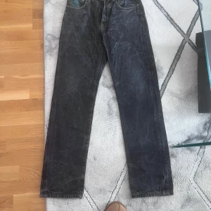 Svarta jeans med broderade detaljer från RMC - Unika svarta jeans från RMC med stora broderade röda tecken på bakfickorna och turkosa detaljer. Jeansen har en knappgylf och klassisk femficksdesign. Perfekta för dig som vill sticka ut med en cool och annorlunda stil. Wasit30 Length 32