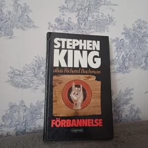 Förbannelse - Boken 'Förbannelse' av Stephen King, skriven under pseudonymen Richard Bachman, är en spännande thriller. Omslaget är svart med en bild av en hand som gör ett tecken. Texten är i vita och röda bokstäver. Perfekt för dig som älskar skräck och mystik!