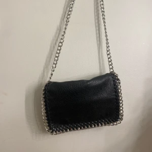 Svart axelväska med kedjedetaljer - Snygg svart axelväska i skinn med coola kedjedetaljer runt kanterna. Väskan har en praktisk dragkedja på baksidan för extra förvaring. Perfekt för att ge din outfit en edgy touch.