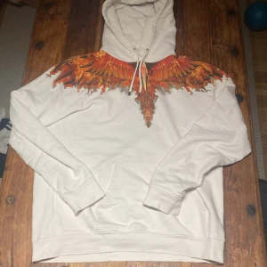 Vit hoodie från Marcelo Burlon - Säljer en as fet vit hoodie från Marcelo Burlon med ett coolt orange och rött vingmönster över axlarna. Hoodien har en normal passform med snören i huvan. Väldigt bra skick. Perfekt för en stilren look! Kom gärna med bud! 