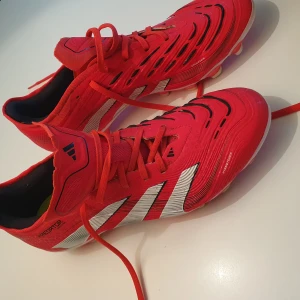 Adidas Predator Pro fg/ag (MG) fotbollsskor  - Röda Adidas Predator Pro MG fotbollsskor med vita och svarta detaljer. Skorna har snörning, rund tå och platt sula med fasta dobbar för spel på gräs och konstgräs. Använda 1 vecka och kändes extremt bra men var fel storlek. 