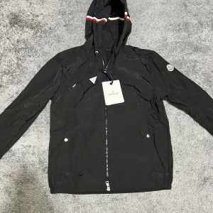 Svart vindjacka från Moncler med huva - Svart vindjacka från Moncler med huva och dragkedja framtill. Jackan har Moncler-logga på ärmen och vita samt röda detaljer på huvan. Två fickor med knappar och justerbar dragsko i huvan. Perfekt för dig som gillar stilren design. Priset kan diskuteras!