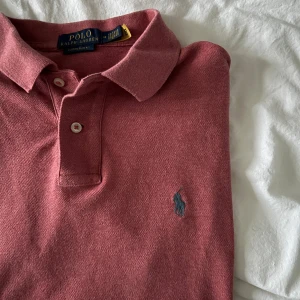 Rosa pikétröja från Ralph Lauren - Säljer en klassisk rosa pikétröja från Ralph Lauren med grå broderad logga på bröstet. Tröjan har korta ärmar, krage och två knappar framtill. Perfekt för en stilren och avslappnad look.