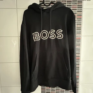 Svart hoodie från BOSS - Svart hoodie från BOSS med stor vit broderad logga på bröstet. Tröjan har huva med dragsko och en stor magficka framtill. Perfekt för en avslappnad och stilren look. 