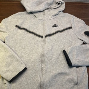 Nike Tech Fleece - Snygg ljusgrå hoodiejacka från Nike med dragkedja och svart logga på bröstet. Jackan har huva, svarta detaljer längs ärmslut och bröst samt praktiska fickor. Perfekt för en sportig och avslappnad stil.
