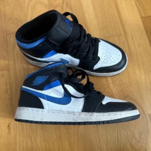 Nike Air Jordans Mid svart/blå/vit - Säljer ett par Nike Air Jordans Mid i svart, blått och vitt. Skorna har klassisk snörning, perforerad tå och den ikoniska Air Jordan-loggan på sidan och plösen. Perfekta för dig som gillar streetwear och sneakers med attityd.