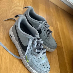 Grå Nike Air Force 1 sneakers - Säljer ett par grå Nike Air Force 1 sneakers med svart swoosh och ljusgröna detaljer. Skorna har klassisk snörning och perforerad tå för extra ventilation. Perfekta för dig som gillar stilrena och sportiga sneakers.